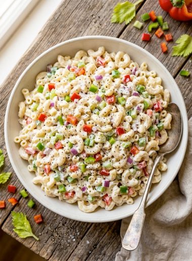 A large white bowl of creamy macaroni salad with red and green bell peppers, celery, and red onion, shot from above on a rustic wooden table
