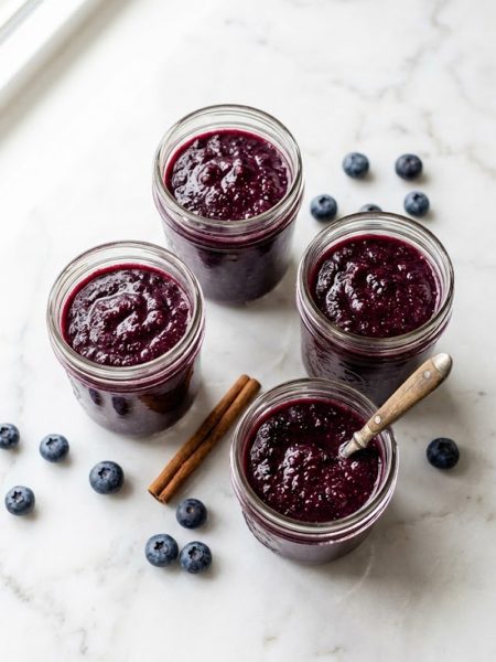 Four glass jars filled with vibrant homemade blueberry applesauce on a white marble surface
