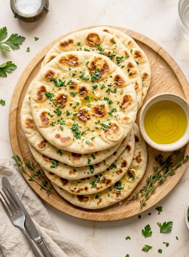 A stack of warm soft homemade Greek pita breads with beautiful golden char spots brushed with herb butter and sprinkled with fresh parsley and flaky sea salt on a wooden board