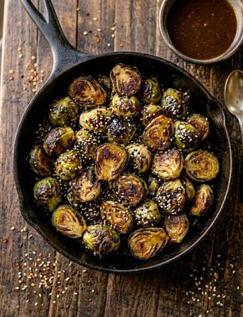 Crispy roasted Brussels sprouts coated in a sticky honey garlic glaze with sesame seeds in a black cast iron skillet on a dark rustic surface