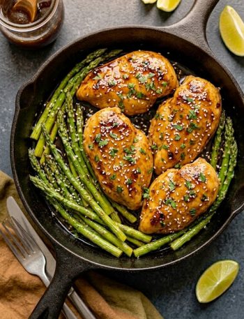 Honey garlic chicken breasts with asparagus in a cast iron skillet, topped with sesame seeds and fresh parsley