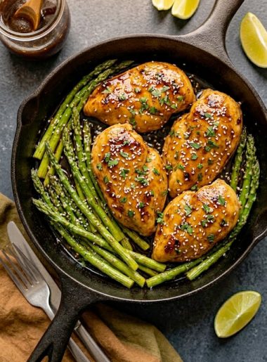 Honey garlic chicken breasts with asparagus in a cast iron skillet, topped with sesame seeds and fresh parsley
