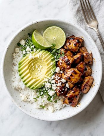 A white ceramic bowl filled with sticky honey lime glazed chicken, fluffy white rice, sliced avocado, crumbled cotija cheese, fresh cilantro, and lime wedges