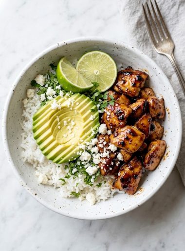 A white ceramic bowl filled with sticky honey lime glazed chicken, fluffy white rice, sliced avocado, crumbled cotija cheese, fresh cilantro, and lime wedges