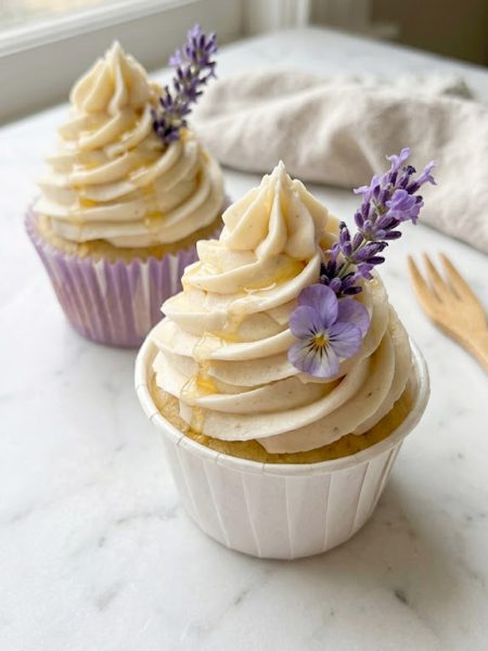 A close-up of two elegant lavender honey cupcakes with tall swirled honey buttercream frosting topped with fresh purple lavender sprigs and edible flowers in white and purple cupcake liners on a white marble surface