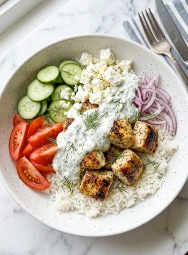 A large white ceramic bowl filled with fluffy white rice topped with golden seared lemon dill chicken chunks fresh cucumber slices ripe tomato wedges thinly sliced red onion crumbled feta cheese and a generous spoonful of creamy dill sauce garnished with fresh dill fronds