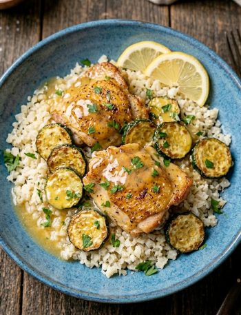 Golden seared lemon butter chicken pieces with crispy zucchini rounds served over cauliflower rice in a wide blue bowl garnished with fresh parsley and lemon slices