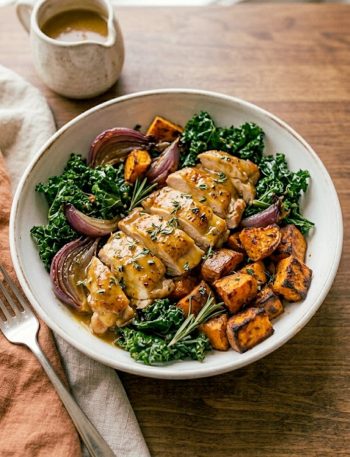 A stunning bowl of maple Dijon chicken with caramelized roasted sweet potatoes wilted kale and roasted red onion drizzled with a golden maple Dijon sauce in a wide ceramic bowl