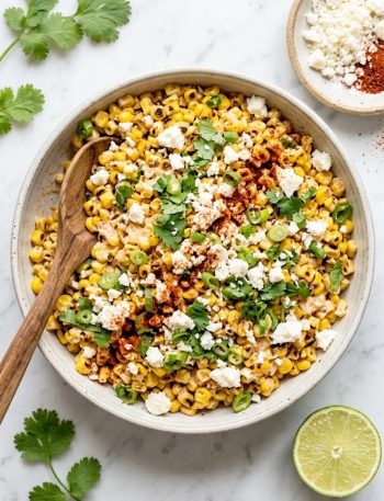 A large ceramic bowl of Mexican street corn salad with golden charred corn kernels, crumbled cotija cheese, fresh cilantro, sliced green onions, and a dusting of chili powder and smoked paprika, shot from above on a light marble surface