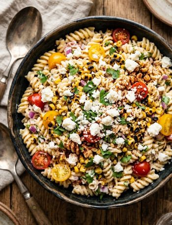 Creamy Mexican street corn pasta salad with rotini pasta sweet corn cherry tomatoes red onion cotija cheese and fresh cilantro in a dark ceramic bowl on a rustic wooden surface