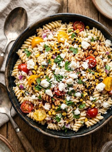 Creamy Mexican street corn pasta salad with rotini pasta sweet corn cherry tomatoes red onion cotija cheese and fresh cilantro in a dark ceramic bowl on a rustic wooden surface