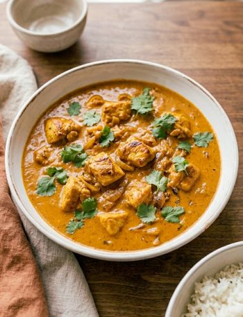 A white bowl of rich creamy Moroccan chicken in a deeply spiced orange tomato sauce garnished with fresh cilantro served alongside fluffy white rice