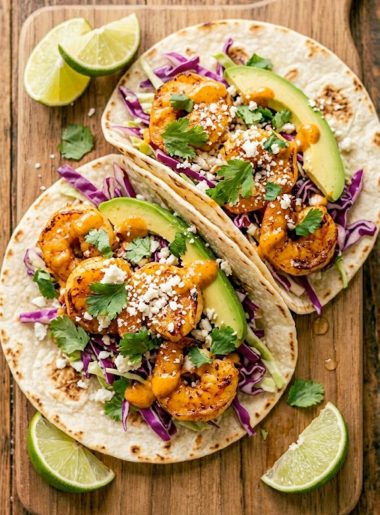 Two mouthwatering shrimp tacos on a wooden board topped with purple cabbage slaw, avocado, crumbled cotija cheese, and chipotle drizzle with lime wedges on the side