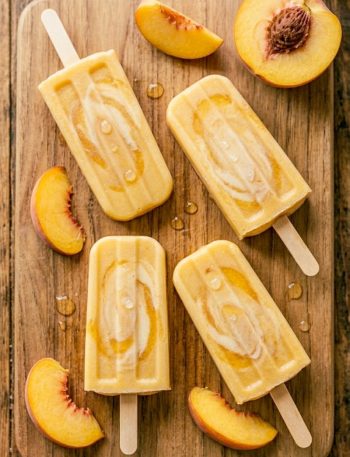 Four golden creamy peaches and cream popsicles on a rustic wooden board surrounded by fresh peach slices with condensation on the surface in warm summer light