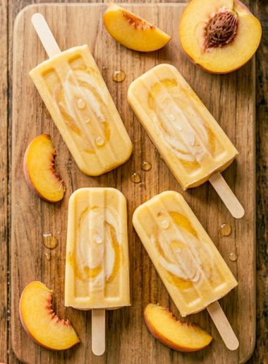 Four golden creamy peaches and cream popsicles on a rustic wooden board surrounded by fresh peach slices with condensation on the surface in warm summer light