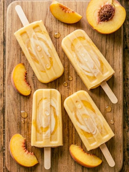 Four golden creamy peaches and cream popsicles on a rustic wooden board surrounded by fresh peach slices with condensation on the surface in warm summer light