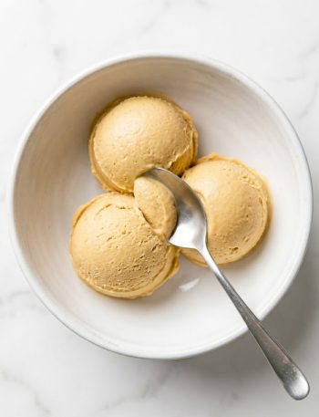 Three scoops of creamy peanut butter cottage cheese ice cream in a white bowl with a silver spoon on a marble surface