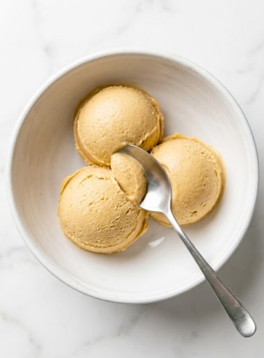 Three scoops of creamy peanut butter cottage cheese ice cream in a white bowl with a silver spoon on a marble surface