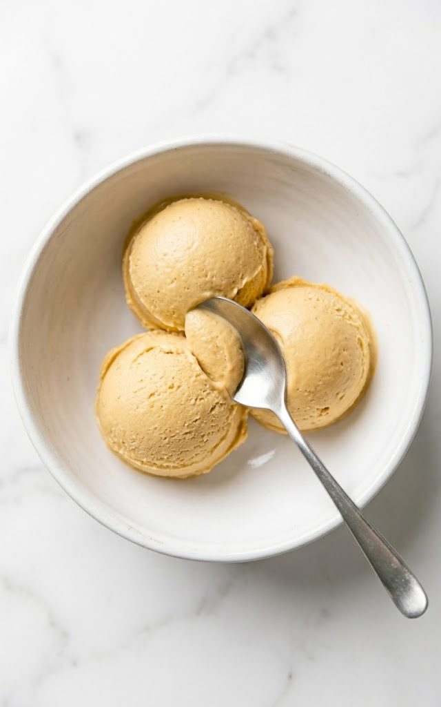 Three scoops of creamy peanut butter cottage cheese ice cream in a white bowl with a silver spoon on a marble surface