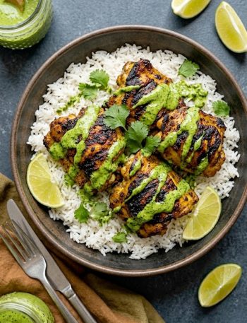 A dark ceramic bowl of smoky charred Peruvian chicken thighs with beautiful char marks served over fluffy white jasmine rice drizzled with bright green cilantro sauce and garnished with fresh cilantro and lime wedges