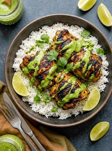 A dark ceramic bowl of smoky charred Peruvian chicken thighs with beautiful char marks served over fluffy white jasmine rice drizzled with bright green cilantro sauce and garnished with fresh cilantro and lime wedges