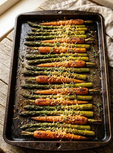 Golden roasted asparagus spears and baby carrots arranged in alternating rows on a dark baking sheet dusted with crispy parmesan and fresh thyme on a rustic wooden surface