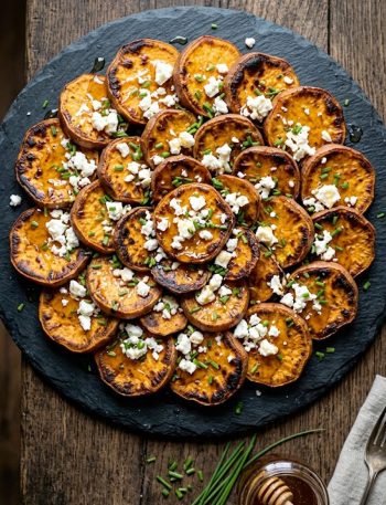 A serving platter of deeply caramelized roasted sweet potato rounds topped with crumbled white feta cheese, a drizzle of golden honey, and freshly chopped chives