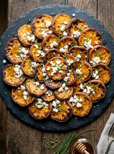 A serving platter of deeply caramelized roasted sweet potato rounds topped with crumbled white feta cheese, a drizzle of golden honey, and freshly chopped chives