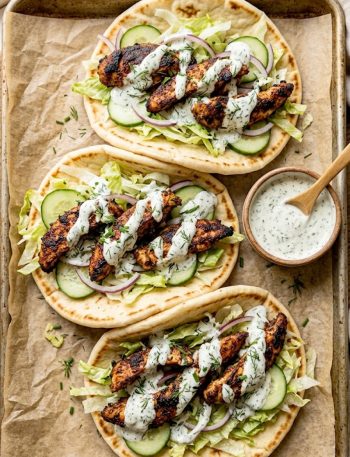 Three sheet pan chicken pitas loaded with smoky spiced chicken strips shredded lettuce sliced cucumber red onion and a generous drizzle of creamy herby ranch sauce on a parchment lined baking sheet