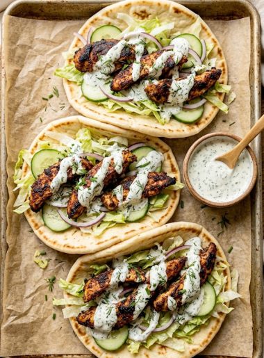 Three sheet pan chicken pitas loaded with smoky spiced chicken strips shredded lettuce sliced cucumber red onion and a generous drizzle of creamy herby ranch sauce on a parchment lined baking sheet