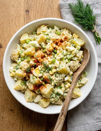 A large white bowl of southern potato salad with tender yellow potatoes, chopped hard boiled eggs, celery, green onions, fresh dill, and a creamy mayo dressing topped with a dusting of smoked paprika, shot from above on a rustic wooden surface