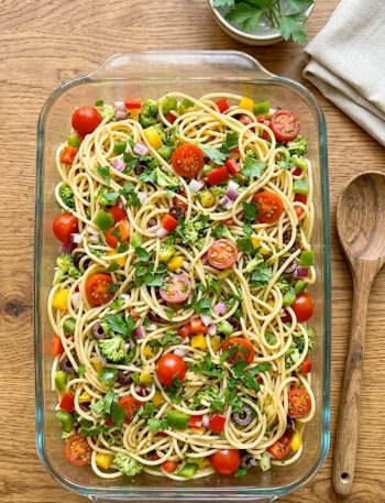 A large glass baking dish filled with cold spaghetti pasta salad loaded with colorful diced red green and yellow bell peppers, tomatoes, broccoli, red onion, black olives and fresh parsley tossed in a zesty Italian dressing, shot from above on a warm wooden surface