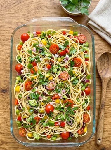 A large glass baking dish filled with cold spaghetti pasta salad loaded with colorful diced red green and yellow bell peppers, tomatoes, broccoli, red onion, black olives and fresh parsley tossed in a zesty Italian dressing, shot from above on a warm wooden surface