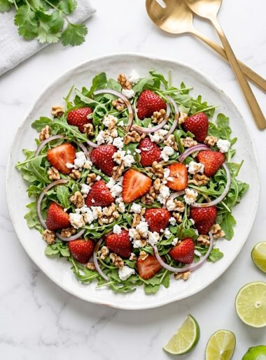 A large white plate of spring strawberry arugula salad with fresh sliced strawberries, peppery arugula leaves, crumbled goat cheese, chopped walnuts, and honey balsamic vinaigrette drizzled on top, shot from above on a bright marble surface