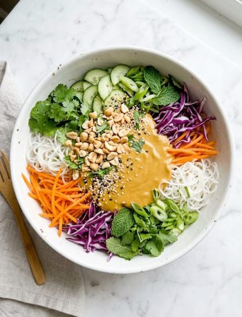 A top-down view of a vibrant spring roll salad with rice vermicelli noodles, shredded orange carrots, purple cabbage, fresh cilantro, mint leaves, and green onions topped with a creamy golden peanut dressing, chopped roasted peanuts, and sesame seeds in a large white bowl on a marble surface