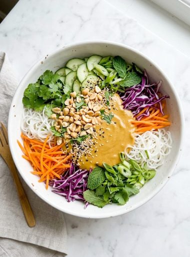 A top-down view of a vibrant spring roll salad with rice vermicelli noodles, shredded orange carrots, purple cabbage, fresh cilantro, mint leaves, and green onions topped with a creamy golden peanut dressing, chopped roasted peanuts, and sesame seeds in a large white bowl on a marble surface