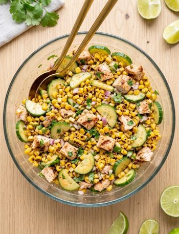 A large glass bowl of street corn creamy cucumber chicken salad with grilled chicken pieces, charred corn, sliced cucumbers, diced red onion, fresh cilantro, and a smoky creamy dressing, shot from above on a light wooden surface
