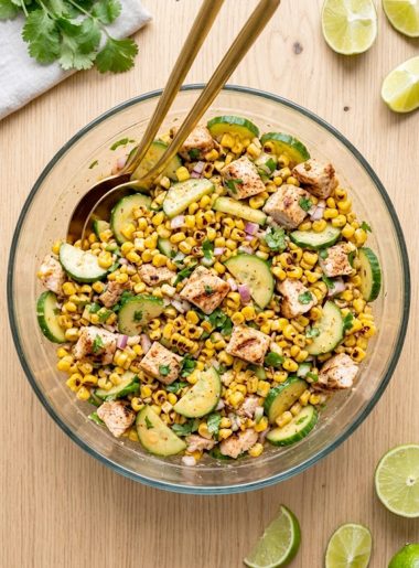 A large glass bowl of street corn creamy cucumber chicken salad with grilled chicken pieces, charred corn, sliced cucumbers, diced red onion, fresh cilantro, and a smoky creamy dressing, shot from above on a light wooden surface