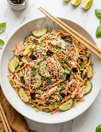 A white bowl of sushi cucumber salad with imitation crab, sliced cucumbers, nori pieces, black sesame seeds and creamy spicy mayo dressing with chopsticks resting on the side