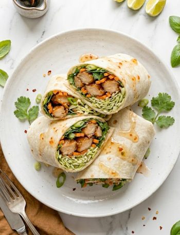 Two sweet chili chicken wraps cut in half and stacked on a white plate showing the glossy glazed chicken crunchy cabbage slaw shredded carrots fresh cilantro and lime wedges