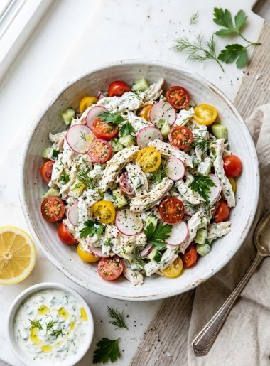 A bowl of creamy tzatziki chicken salad with visible cherry tomatoes, radishes, cucumber, and fresh dill, served on a light wooden surface