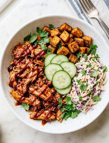 A wide white ceramic bowl filled with saucy shredded BBQ chicken, crispy roasted sweet potato cubes, creamy purple and green coleslaw, and fresh cucumber rounds topped with parsley