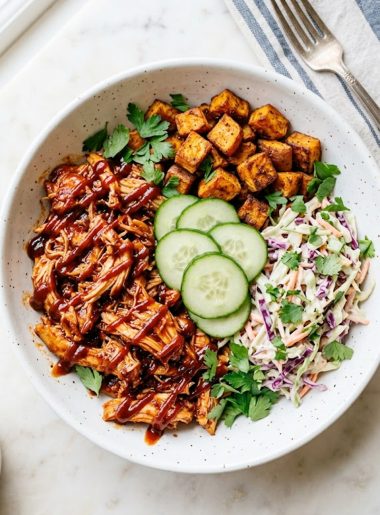 A wide white ceramic bowl filled with saucy shredded BBQ chicken, crispy roasted sweet potato cubes, creamy purple and green coleslaw, and fresh cucumber rounds topped with parsley