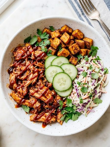 A wide white ceramic bowl filled with saucy shredded BBQ chicken, crispy roasted sweet potato cubes, creamy purple and green coleslaw, and fresh cucumber rounds topped with parsley