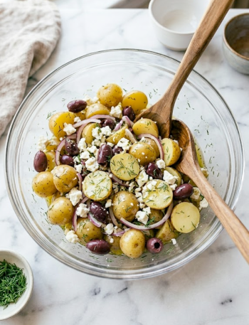 A glass bowl of Greek olive potato salad with baby yellow potatoes, Kalamata olives, crumbled feta, red onion, and fresh dill on a marble surface