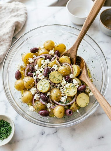 A glass bowl of Greek olive potato salad with baby yellow potatoes, Kalamata olives, crumbled feta, red onion, and fresh dill on a marble surface