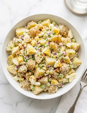 A large white ceramic bowl of creamy all-American potato salad made with tender Yukon Gold potatoes, hard-boiled eggs, red onion, celery, and fresh chives topped with a dusting of smoked paprika on a white marble surface