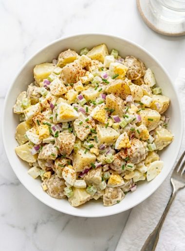 A large white ceramic bowl of creamy all-American potato salad made with tender Yukon Gold potatoes, hard-boiled eggs, red onion, celery, and fresh chives topped with a dusting of smoked paprika on a white marble surface