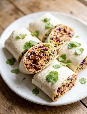 Four BBQ chicken coleslaw wraps cut in half and arranged on a white plate, showing smoky shredded chicken and creamy coleslaw filling, garnished with fresh cilantro