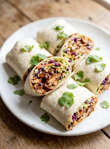 Four BBQ chicken coleslaw wraps cut in half and arranged on a white plate, showing smoky shredded chicken and creamy coleslaw filling, garnished with fresh cilantro
